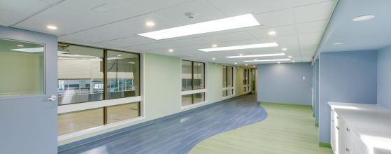 A brightly lit hallway with blue and green flooring, large windows on one side, overhead lights, and white cabinets along the wall.